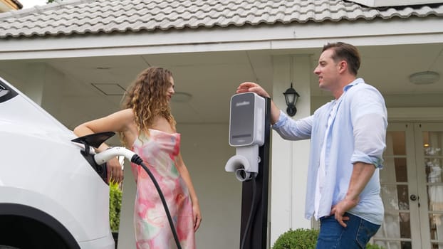 Happy and lovely couple with eco-friendly conscious recharging electric vehicle from EV charging station. EV car technology utilized as alternative transportation for future sustainability. Synchronos