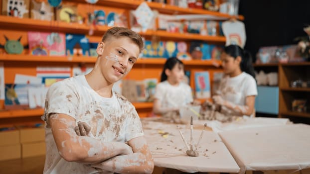Smiling boy looking at camera and crossing arm with confident at workshop while diverse student having pottery class together. Happy caucasian student smile while pose with arm folded. Edification.