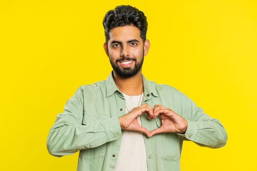 Man in love. Smiling attractive Indian man makes heart gesture demonstrates love sign expresses good positive feelings and sympathy. Handsome Arabian Hindu young guy isolated on yellow wall background