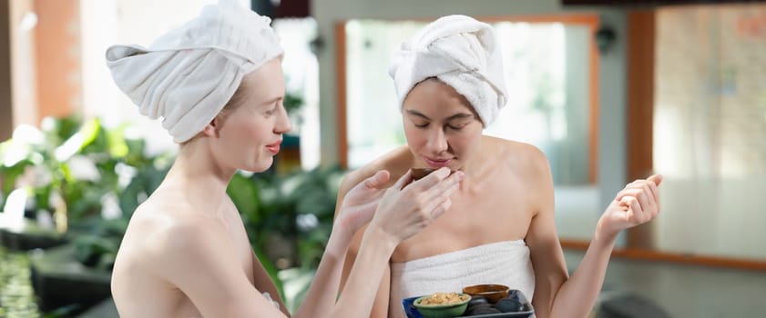 Couple of beautiful woman interested in homemade facial masks while sitting at spa salon. Attractive woman in white towel enjoy herbal masks with her friend surrounded by nature. Tranquility.