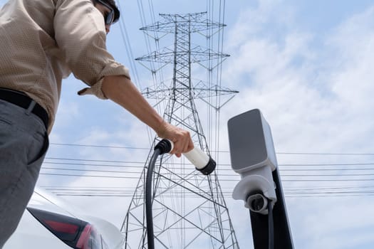 Man recharge EV electric car battery at charging station connected to electrical power grid tower on sky background as electrical industry for eco friendly vehicle utilization. Expedient