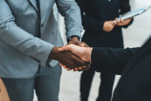 Closeup of business hands shaking between businessman and professional male leader while smart businesswoman stand near at modern office corridor. Represented unity, corporate, teamwork. Ornamented.