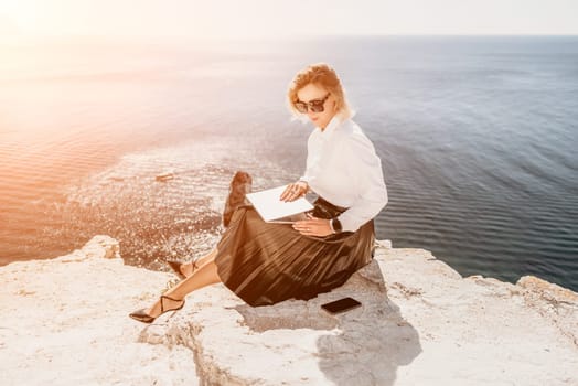 Successful business woman in yellow hat working on laptop by the sea. Pretty lady typing on computer at summer day outdoors. Freelance, travel and holidays concept.