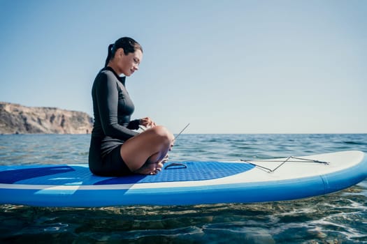 Digital nomad, woman in the hat, a business woman with a laptop sits on the rocks by the sea during sunset, makes a business transaction online from a distance. Freelance, remote work on vacation.