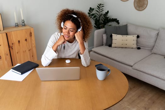Young African American woman working at home office using wireless headphones. Happy Hispanic female entrepreneur using laptop. Working at home concept.