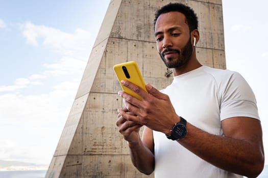 African American man in sports clothes checking fitness app on mobile phone, using phone to put on music for workout outdoors. Copy space. Sport and technology concept.