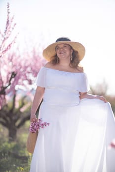 Woman blooming peach orchard. Against the backdrop of a picturesque peach orchard, a woman in a long white dress and hat enjoys a peaceful walk in the park, surrounded by the beauty of nature