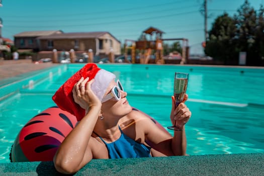 Woman pool Santa hat. A happy woman in a blue bikini, a red and white Santa hat and sunglasses poses near the pool with a glass of champagne standing nearby. Christmas holidays concept