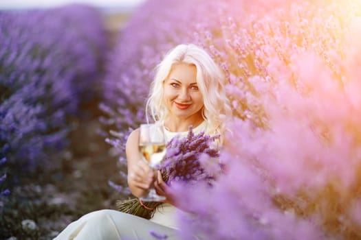 Blonde lavender field holds a glass of white wine in her hands. Happy woman in white dress enjoys lavender field picnic holding a large bouquet of lavender in her hands . Illustrating woman's picnic in a lavender field