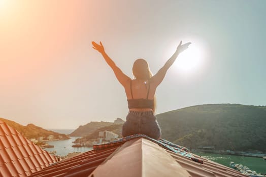 Woman sits on rooftop with outstretched arms, enjoys town view and sea mountains. Peaceful rooftop relaxation. Below her, there is a town with several boats visible in the water.