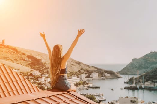 Woman sits on rooftop with outstretched arms, enjoys town view and sea mountains. Peaceful rooftop relaxation. Below her, there is a town with several boats visible in the water.