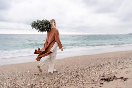 Blond woman Christmas sea. Christmas portrait of a happy woman walking along the beach and holding a Christmas tree on her shoulder. She is wearing a brown coat and a white suit