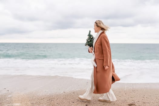 Blond woman Christmas sea. Christmas portrait of a happy woman walking along the beach and holding a Christmas tree in her hands. She is wearing a brown coat and a white suit