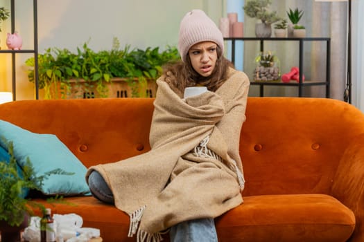 Sick child girl wear hat wrapped in plaid sit alone shivering from cold on sofa drinking hot tea in unheated home apartment without heating due debt. Unhealthy kid feeling discomfort try to warming up