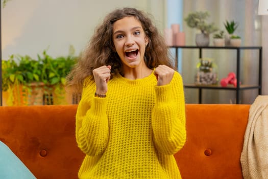 Happy child girl shouting, celebrating success, winning, goal achievement good news, lottery luck victory, watching TV championship at home apartment. Teen 14-15 years kid in living room sits on couch
