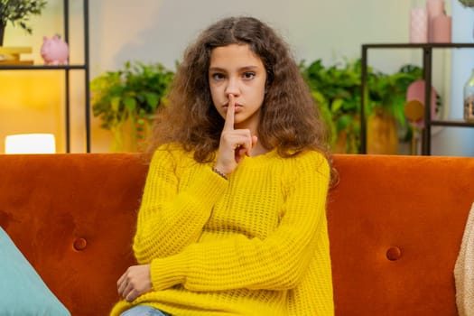 Shh be quiet please. Child girl presses index finger to lips makes silence hush gesture sign do not tell gossip secret. Teenager female 14-15 years kid at home apartment living room sits on couch