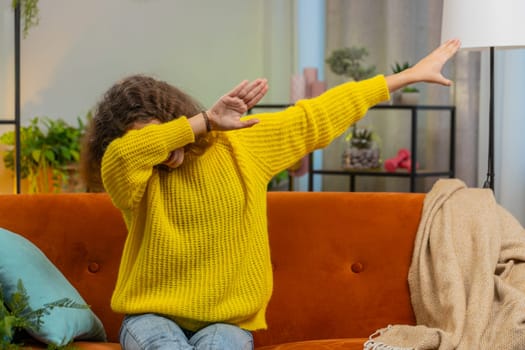 Trendy cheerful positive 14-15 years girl having fun dancing, moving to rhythm dabbing raising hands making dub dance gesture. Teenager Caucasian child kid at home in play living room sitting on couch