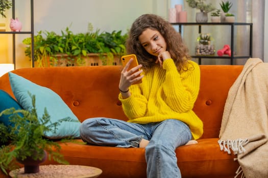 Young teenager child girl texting share messages content on smartphone social media applications online watching relax movie. Caucasian smiling kid uses mobile phone at home in living room sit on sofa