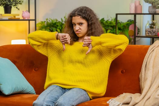 Dislike. Upset child girl showing thumbs down sign gesture, expressing discontent, disapproval, dissatisfied bad work at modern home apartment indoors. Displeased teenager 14-15 years kid on room sofa