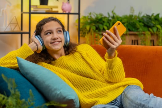 Happy excited overjoyed teenager girl kid in wireless headphones relaxing lying on couch at home room choosing listening favorite energetic disco dancing music. Children weekend leisure activities