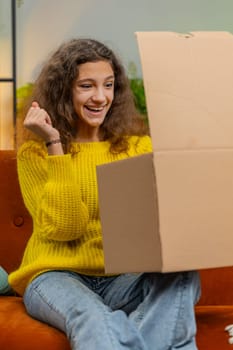 Happy young child girl unpacking delivery parcel. Smiling satisfied teenager kid shopper, online shop customer opening cardboard box receiving purchase gift by fast postal shipping at home. Vertical