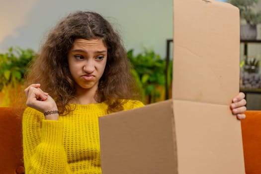 Angry dissatisfied shopper child girl unpacking parcel feeling upset and confused with wrong mistake delivery from an online store, bad quality broken purchase at home. Kid indoors in room on sofa