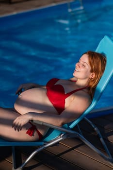 Red-haired pregnant woman in a red bikini sunbathes lying on a sun lounger next to the pool