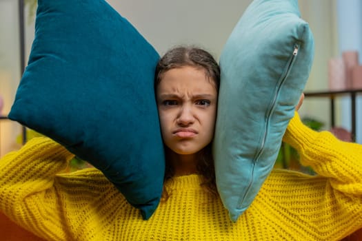 Repair work at neighbors. Irritated angry sad child girl kid cover ears with pillows annoyed by noisy loud music suffers headache wishes silence. Thin walls at home flat without sound insulation