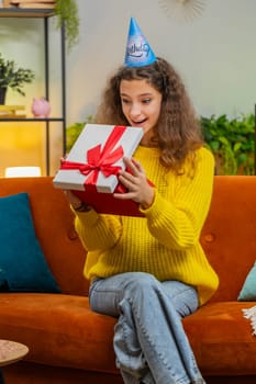Excited happy 14-15 years child girl wears festive birthday cap hat hold gift box with ribbon congratulating. Teenager kid celebrating party event opening delivery greeting present at home. Vertical