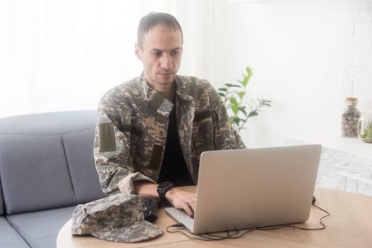 Soldier in office. Serious young military man typing on laptop while working in headquarters building. Caucasian man in camouflage uniform is sitting in bright room at table with notebook and cap. High quality photo