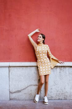 Full body of young female model in stylish, short gown and sneakers looking away while leaning on retro concrete wall with dark red and white colors on street