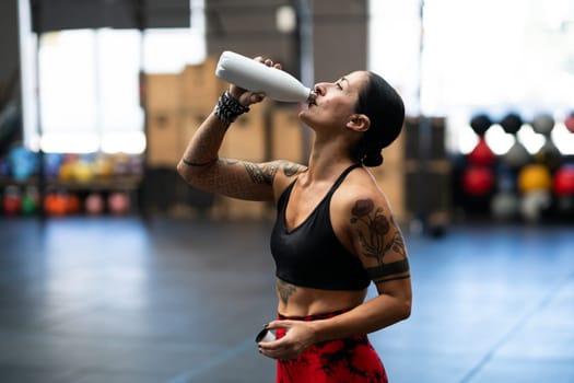 Tired strong mature sportswoman drinking water from a flask in a gym