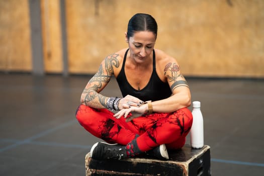 Strong mature sportswoman using smart watch sitting on a box in the gym