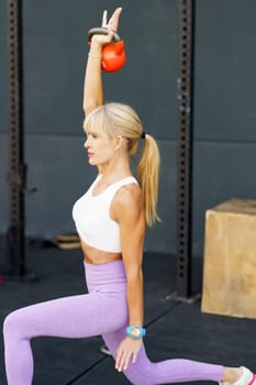 Side view of fit strong female doing kneeling kettlebell press exercise during fitness workout in modern gym against gray wall on floor