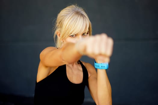 Strong blonde haired female fighter in black tank top in dark gym and practicing punches during martial art, training workout on gray background looking at camera
