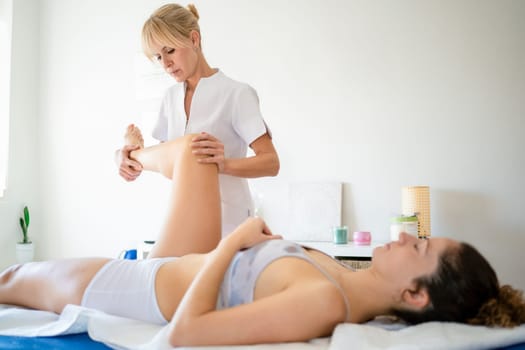 Professional physiotherapist in white uniform doing manipulations on leg of female patient lying on couch during physiotherapy