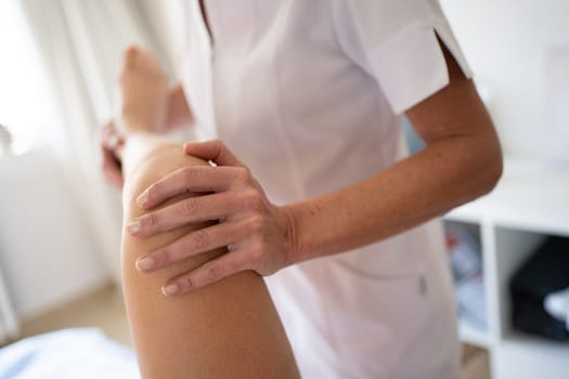 Crop anonymous female physiotherapist in medical uniform doing knee manipulation while treating leg of patient during physiotherapy session