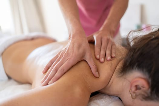 Crop anonymous female massage therapist massaging shoulder of woman during physiotherapy session in light hospital room lying on couch covered with towel