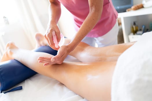 Crop anonymous massage therapist using oily cream while massaging legs of patient lying on stomach during physiotherapy session in modern clinic