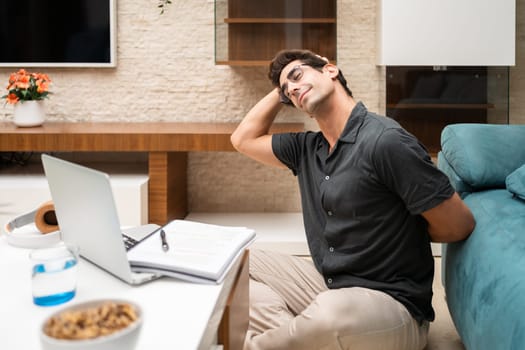 Happy young male freelancer in casual clothes stretching neck while sitting near laptop and notebook in living room at home