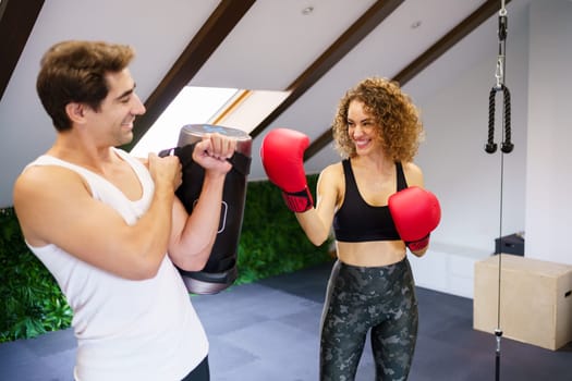 Smiling sporty female boxer in red boxing gloves practicing punches with help of man holding punching bag in gym during training