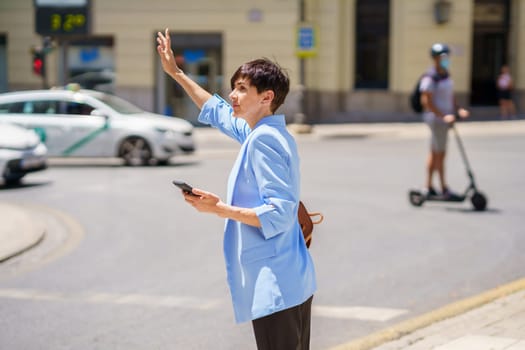 Side view of serious female in formal suit with smartphone at hand standing on city street and catching cab on street