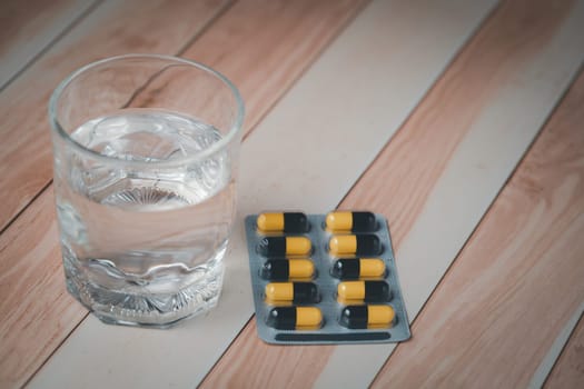 A glass of water and a blister pack of pills, essential for medication, rest on a wooden table, indicating health care routine or treatment