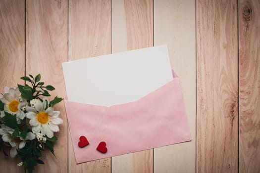 A pink envelope and a white card with a concept of love and Valentine's Day, featuring a red heart and daisy, are gracefully arranged on a wooden table
