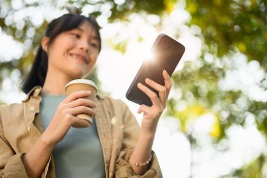 Happy young female freelancer holding paper cup and using smartphone in the summer park.