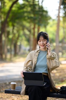 Portrait of smiling young woman talking on mobile phone and working on digital tablet in the park.