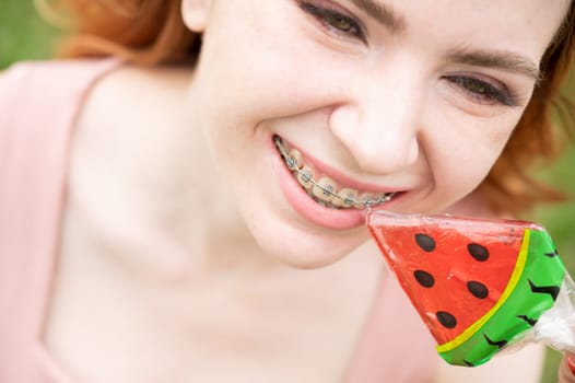 Beautiful young woman with braces on her teeth eats a watermelon-shaped lollipop outdoors