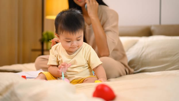 Cute little baby boy playing with toys on bed near working mom. Motherhood and child care concept.