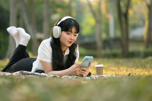Pretty Asian girl with listening music with headphones and using mobile phone on grass.