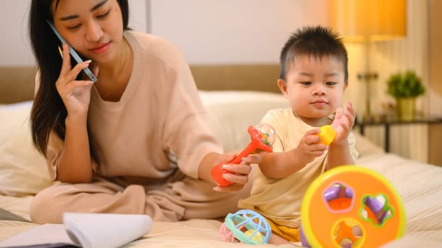 Cute little baby boy playing with toys on bed near working mom. Motherhood and child care concept.